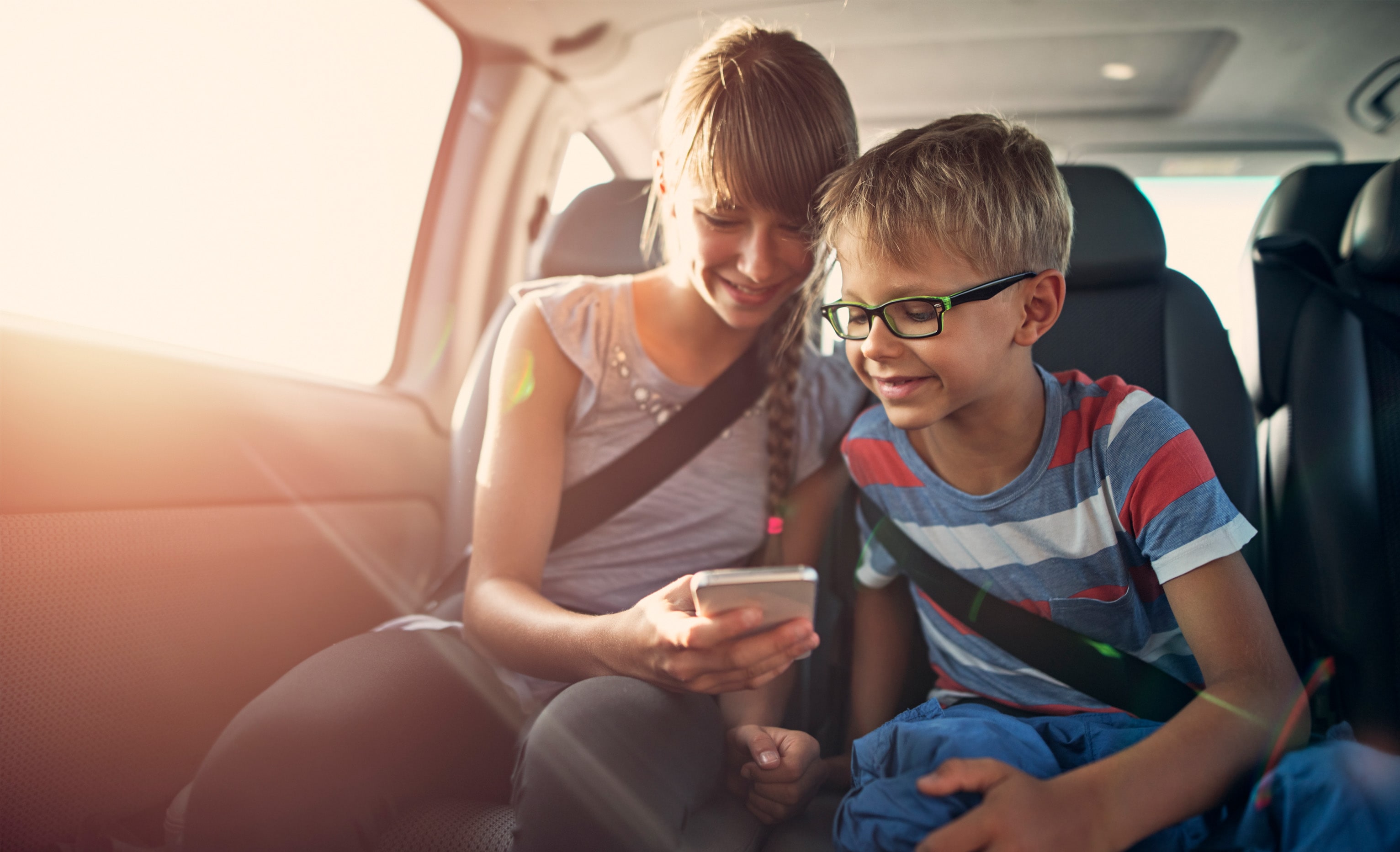 teenage white girl showing mobile phone to a white male child wearing glasses, both smiling and sitting in the back of a car